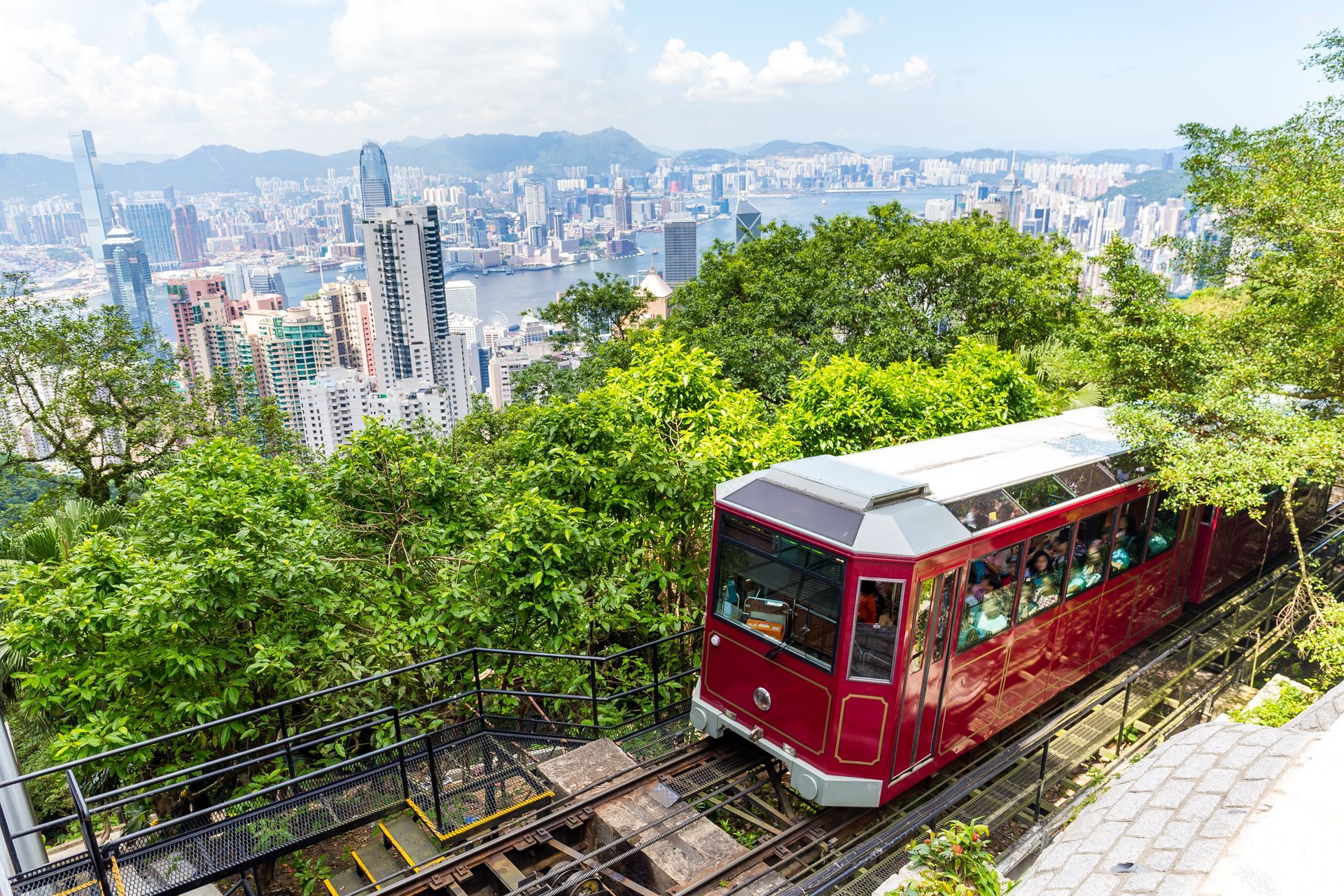 A red tram ascends a steep track on Hong Kong's Peak. Lush green trees frame the city skyline and harbor view.