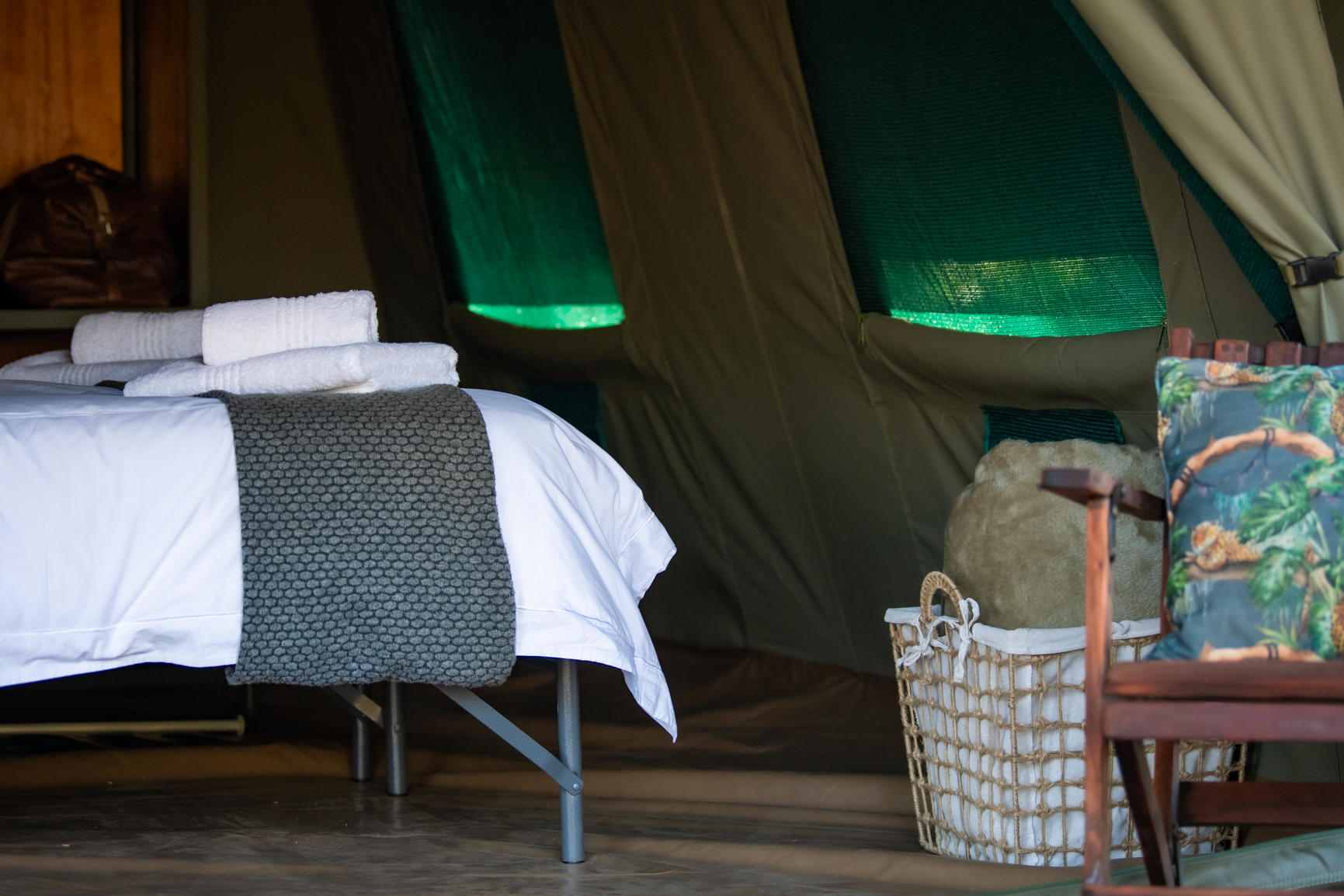 Inside a tent: bed with white linens, gray throw, white towels, wicker basket, wooden chair, and green-lit windows.