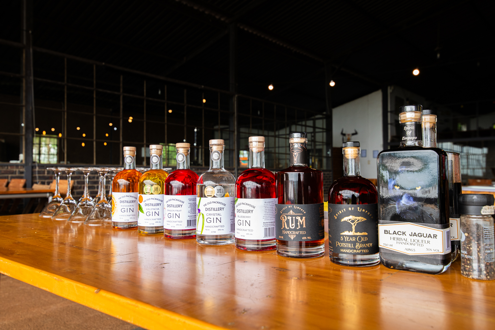 Bottles of liquor in a row on a wooden bar, various colors, glasses to the left, open-air setting.