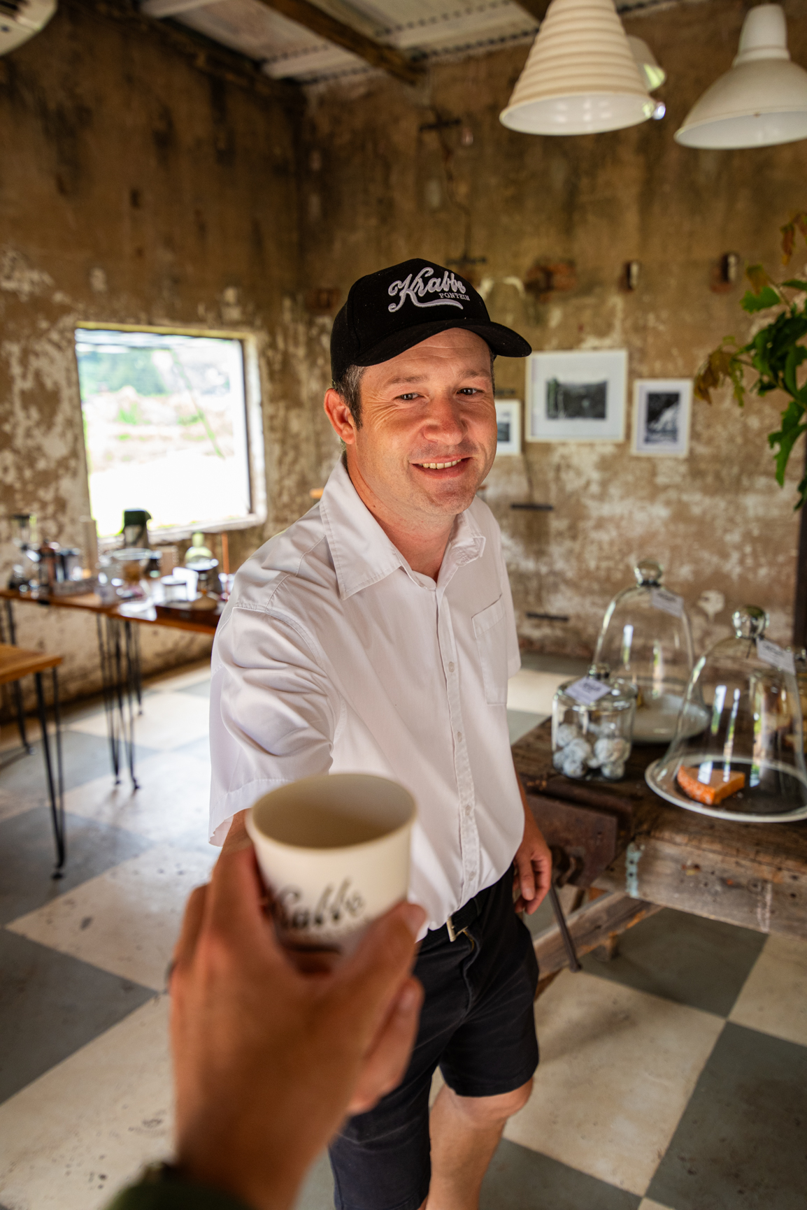 Man in hat smiles, offering a coffee cup in a rustic cafe.