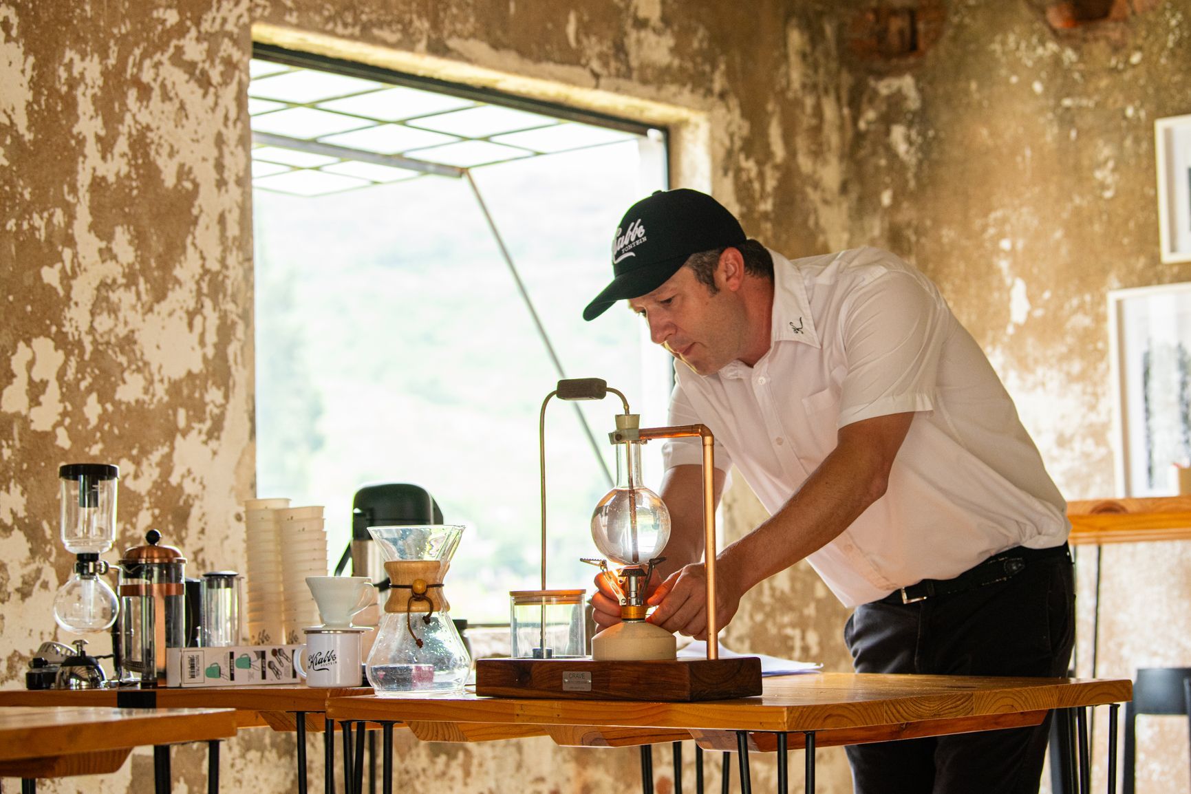 Barista preparing coffee with a siphon brewer at a cafe, wearing a black cap and white shirt.