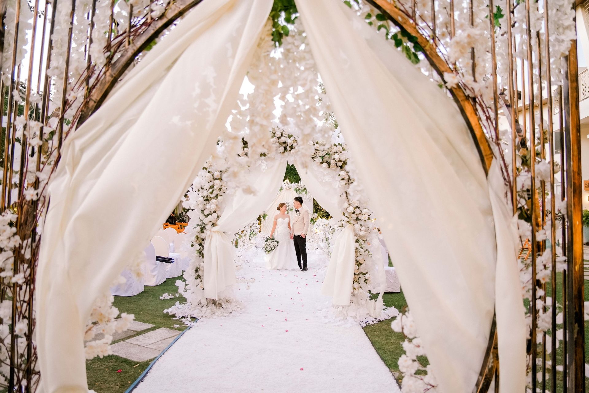 Wedding aisle with couple walking under a floral archway draped in white fabric.