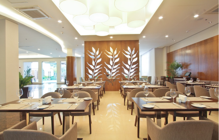 Modern restaurant dining area with tables set for service, warm wood walls, leaf-pattern wall art, and ambient lighting.