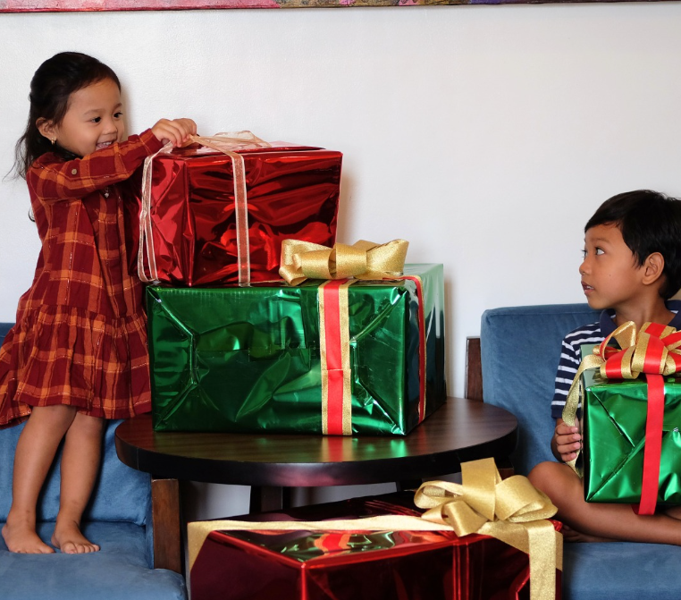 Two children with Christmas gifts. One opens a red gift, the other holds a green one, gold bows.