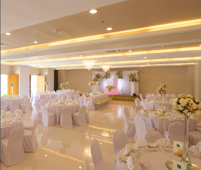 Elegant banquet hall with white tablecloths, chairs, and floral centerpieces; stage backdrop.