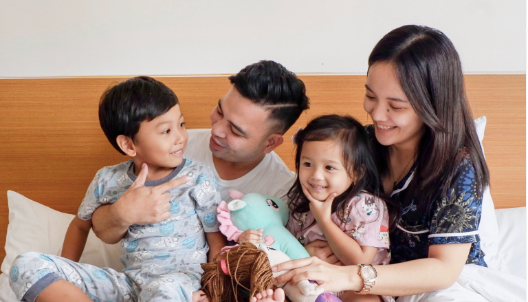 Family sitting on a bed, smiling and playing with a toy and doll in a cozy bedroom.