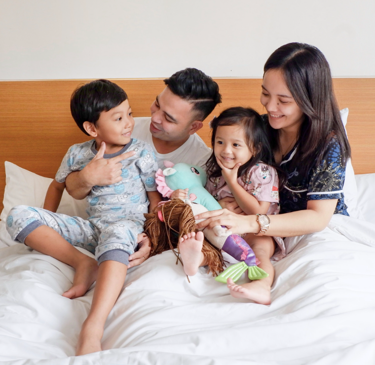 Family of four on a bed, smiling. Boy points, girl holds toy, parents look on.