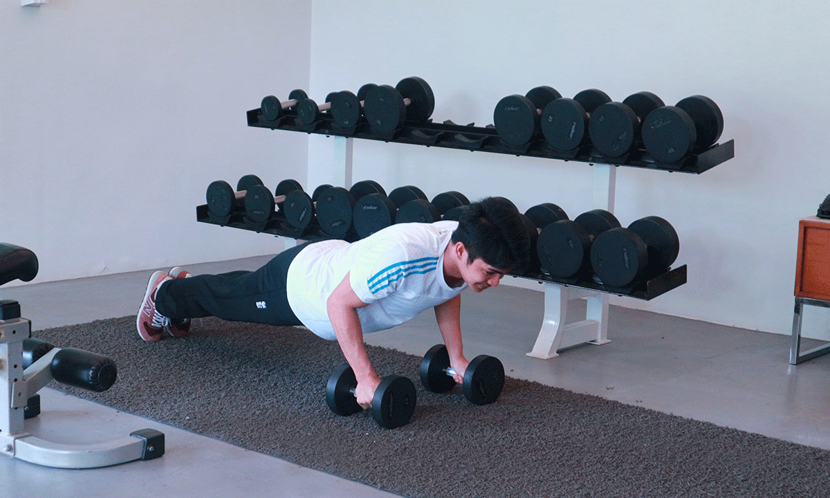 Man doing push-ups on dumbbells in a gym. Black weights on racks in the background.