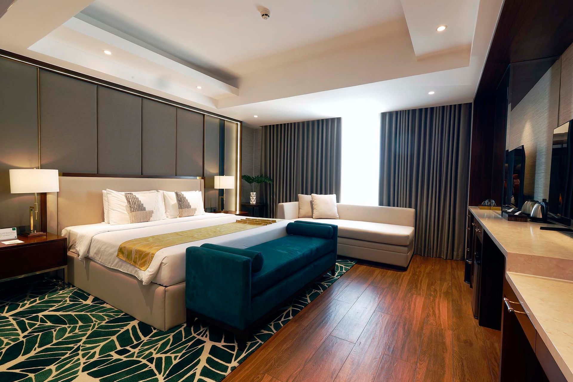 Modern hotel room with king bed, teal bench, wood floor, and dark curtains