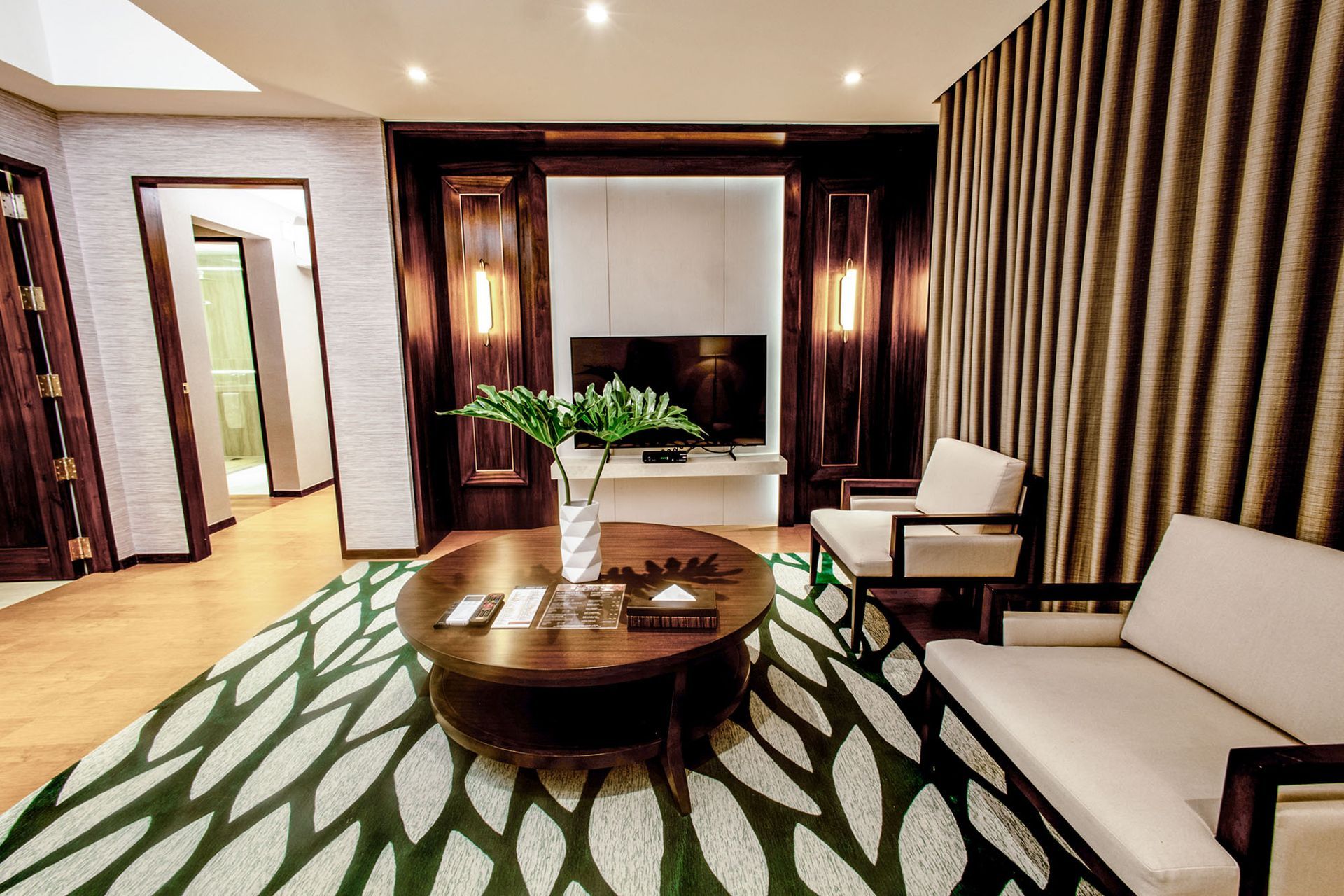 Modern living room with white sofas, round wooden table, green patterned rug, and large window curtains