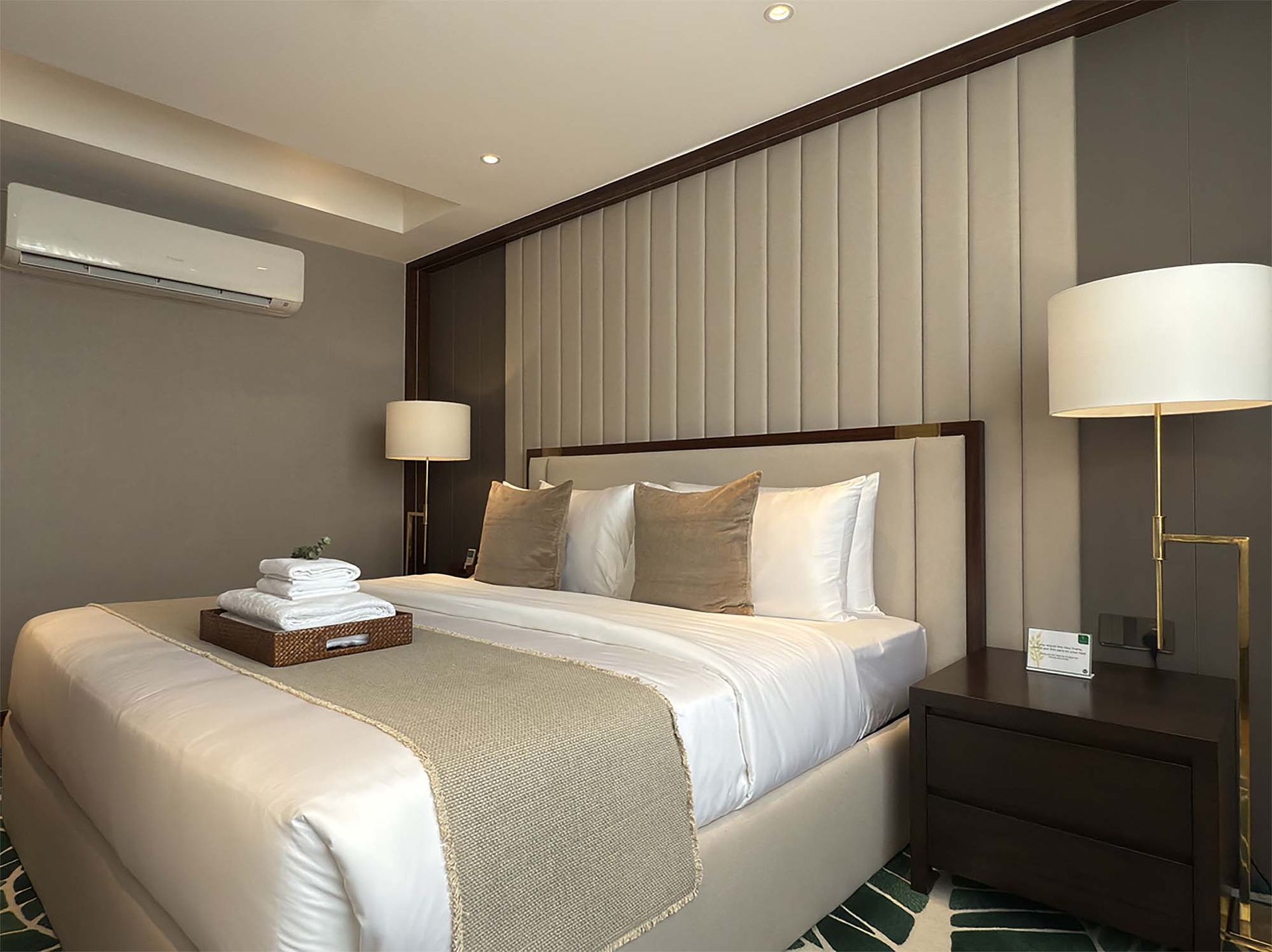 Bedroom with a king-sized bed, beige headboard, and bedside lamps. White linens and a tray of towels sit on the bed.