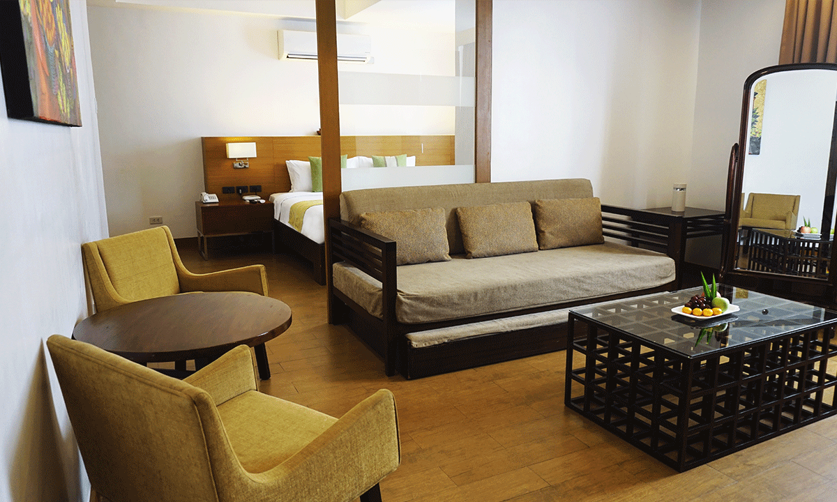 Hotel room with seating area, bed in background, and full-length mirror. Wooden furniture and light wood flooring.
