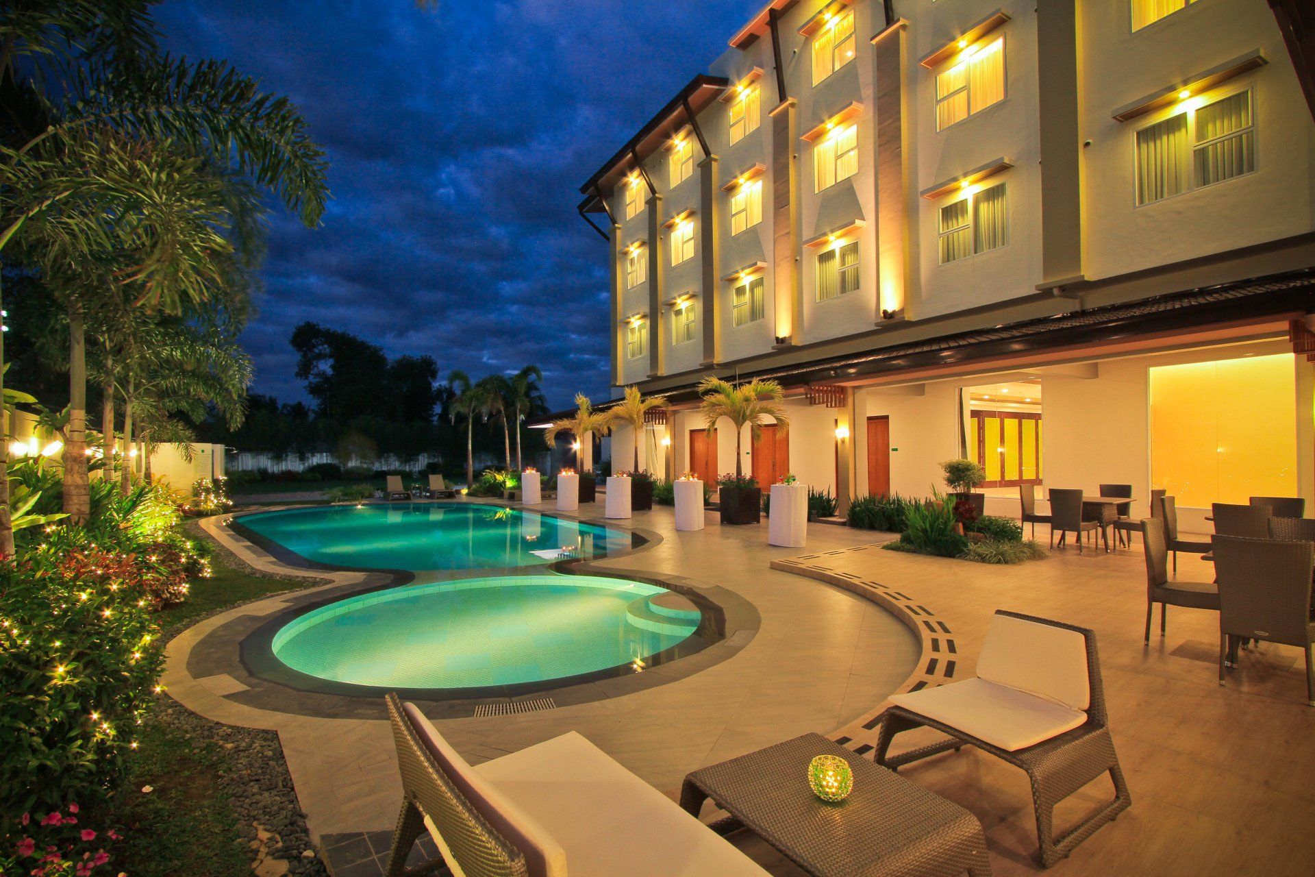 Lit poolside hotel courtyard at dusk with lounge chairs and warm balcony lights