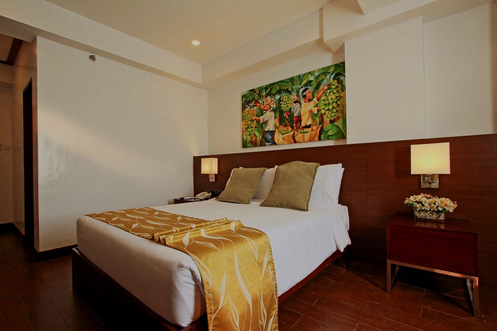 A hotel room featuring a bed with a gold runner, a wooden headboard, two lamps, and a painting on a white wall.