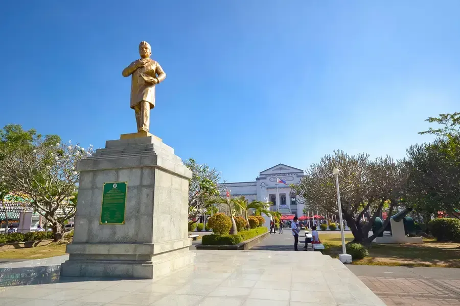 Golden statue on a pedestal in a sunny park with a white building in the background