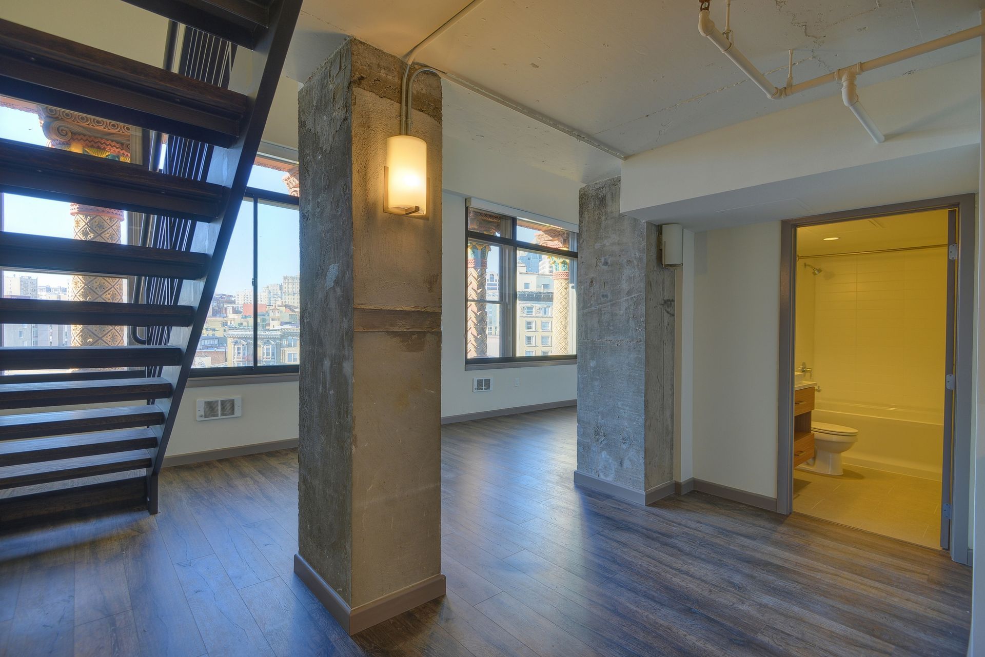 Modern loft interior with staircase, large windows, wood floors, and a lit yellow bathroom doorway