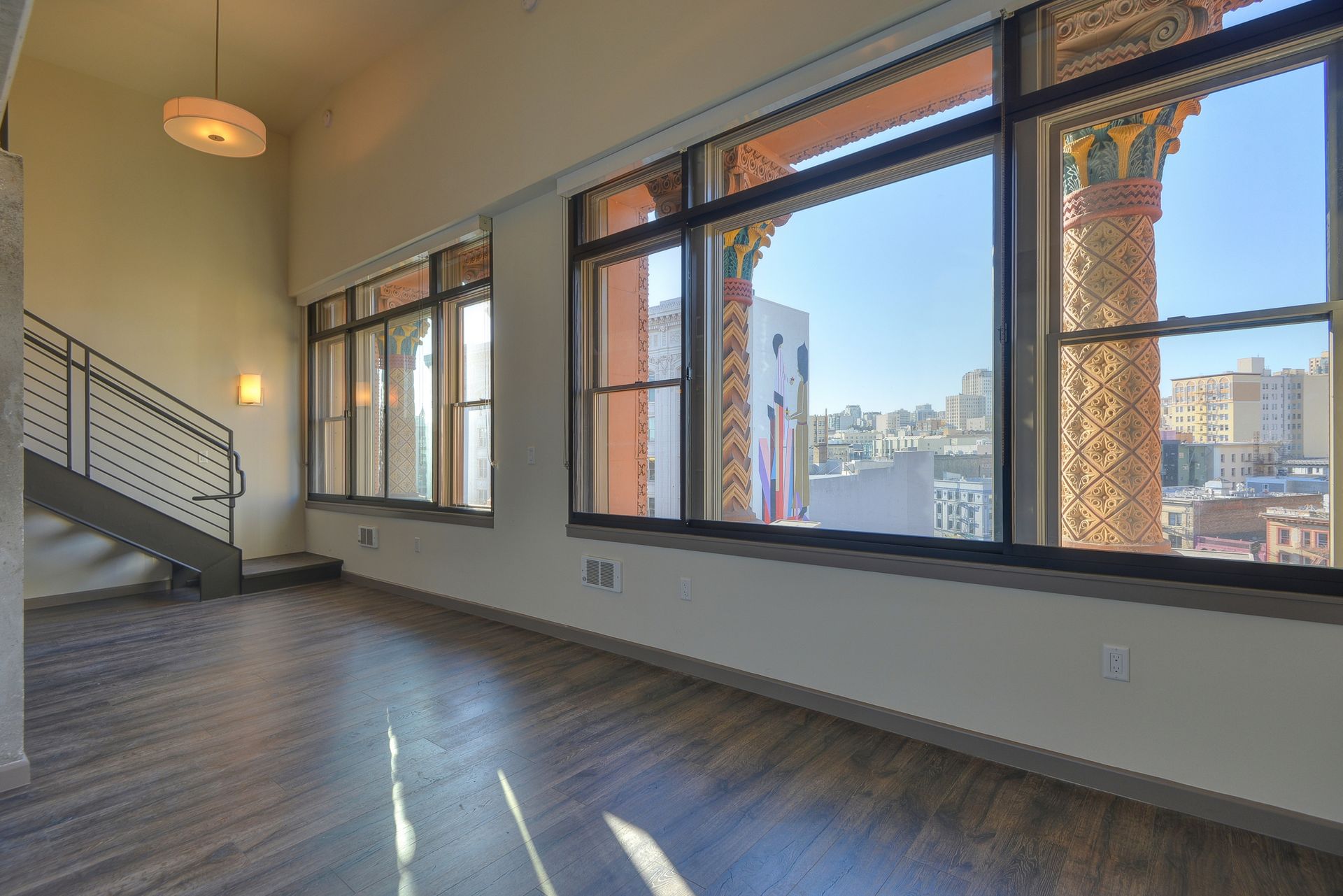 Empty loft-style living room with large windows, hardwood floors, and staircase overlooking city view