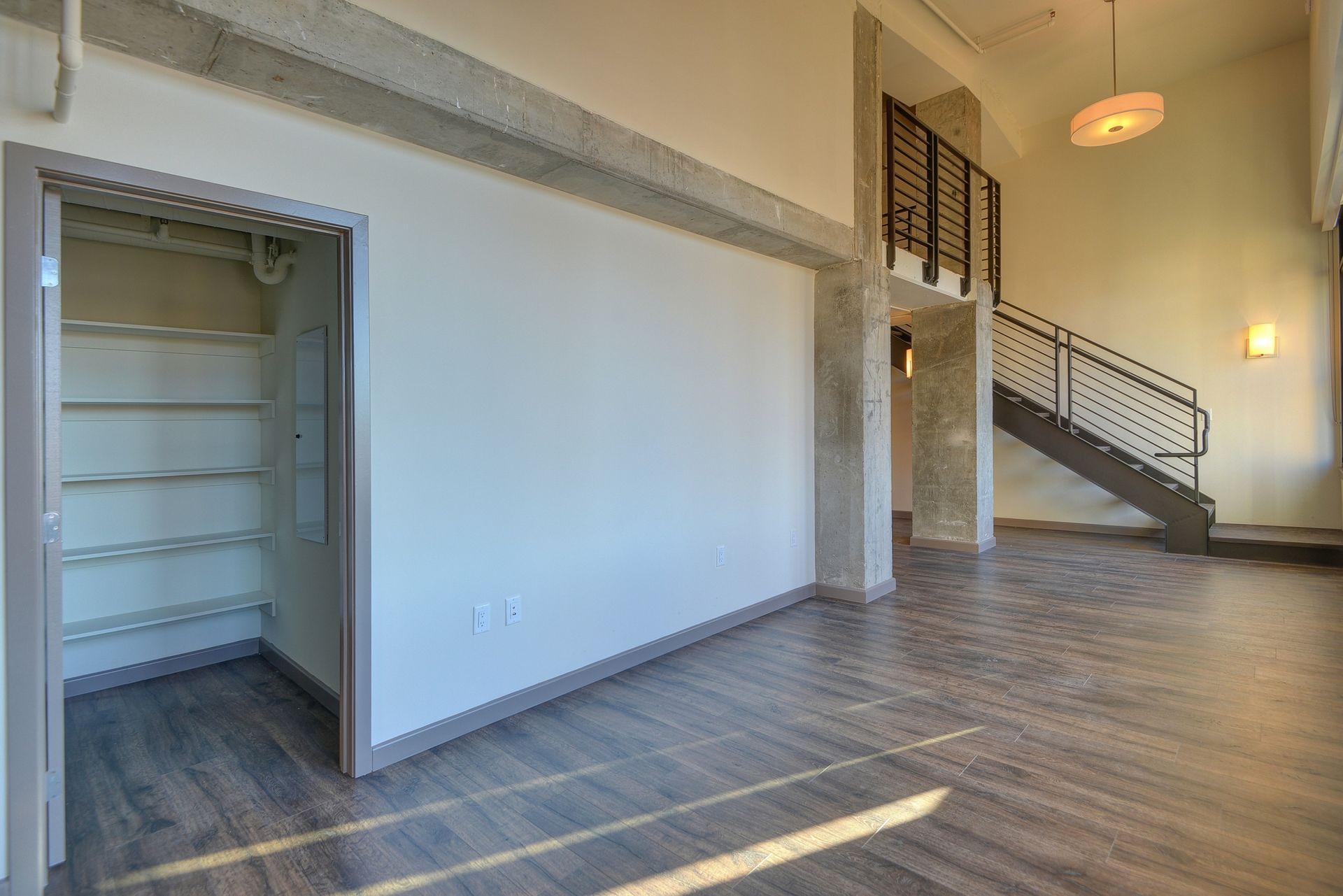 Empty loft with exposed beams, wood floors, tall windows, and a staircase with black railings.