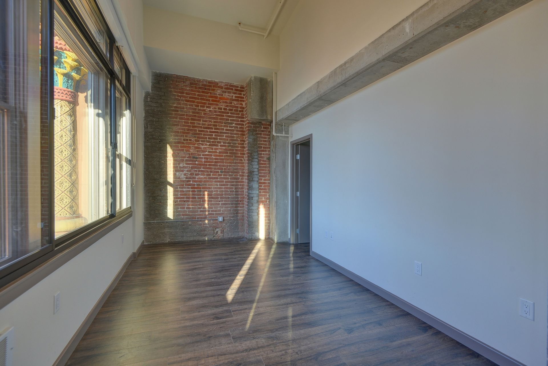 Empty modern room with large windows, exposed brick wall, and sunlight on dark wood floors