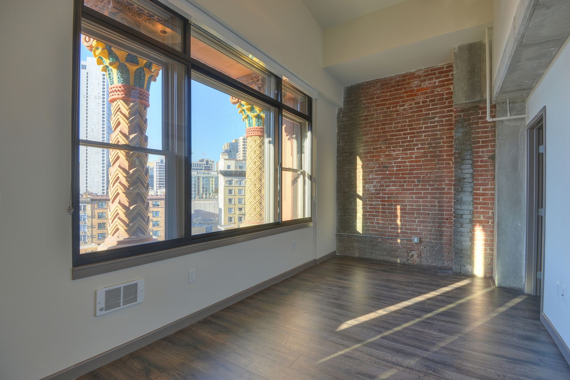 Empty sunlit room with large windows, exposed brick, and herringbone wood flooring