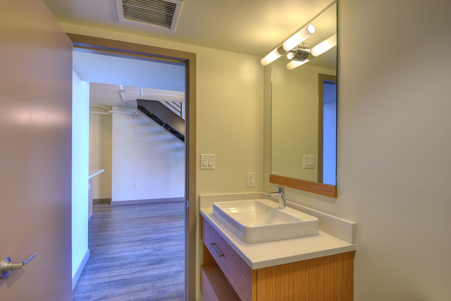 Bathroom vanity and sink with mirror, looking into a room with wood floors and stairs beyond the doorway