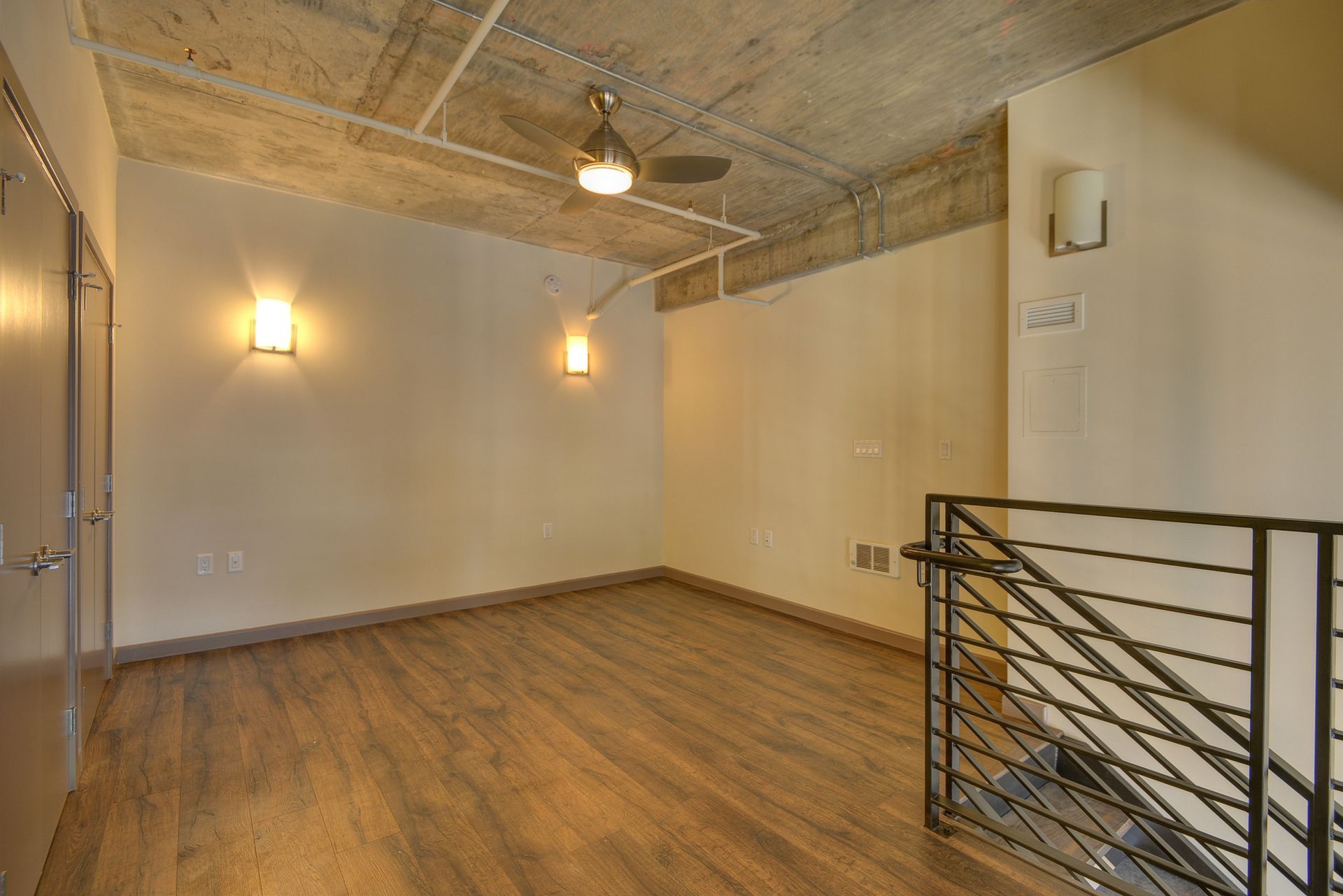 Empty loft with wood floors, exposed concrete ceiling, wall sconces, and metal railing overlooking stairs