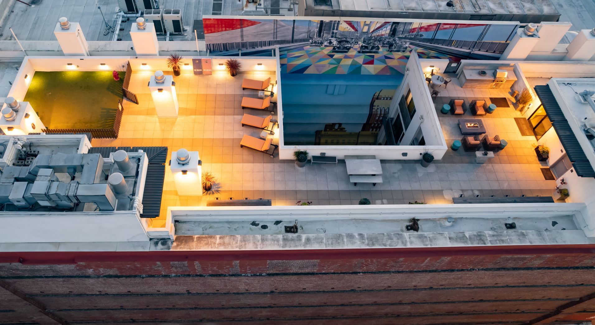 Aerial view of a rooftop deck with lounge chairs, tables, and warm lighting beside a large blue pool.