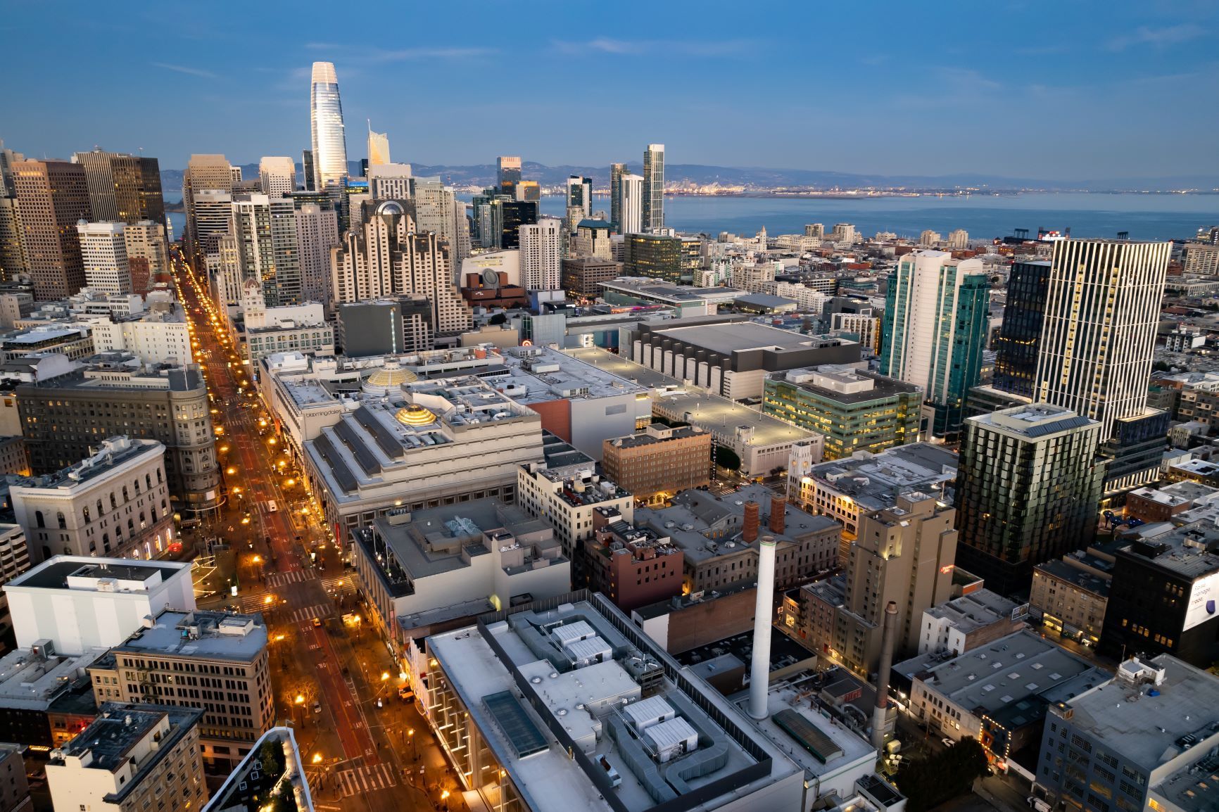 Aerial view of a city skyline at dusk with lit streets and waterfront beyond.