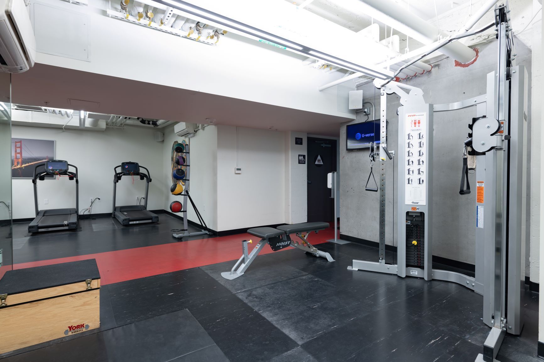 Empty gym with treadmills, cable machine, and weight bench on red and black flooring.