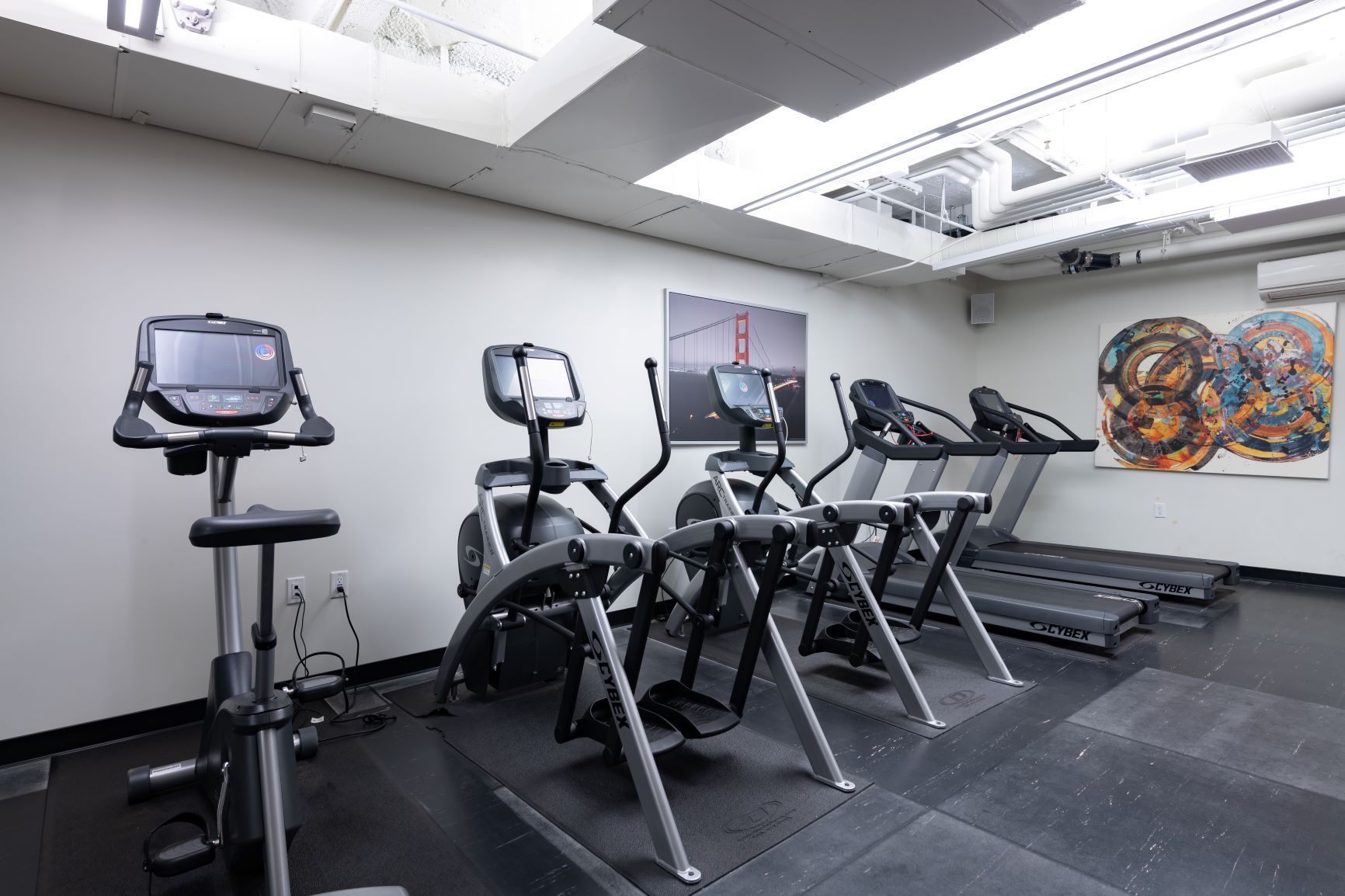 Indoor gym with several treadmills and ellipticals on a rubber floor beneath bright ceiling lights