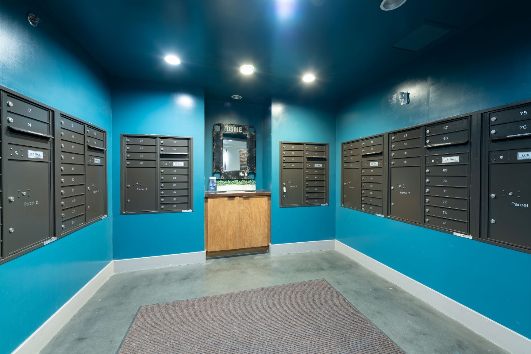 Blue mailroom with rows of wall-mounted mailboxes and a wooden counter under ceiling lights.