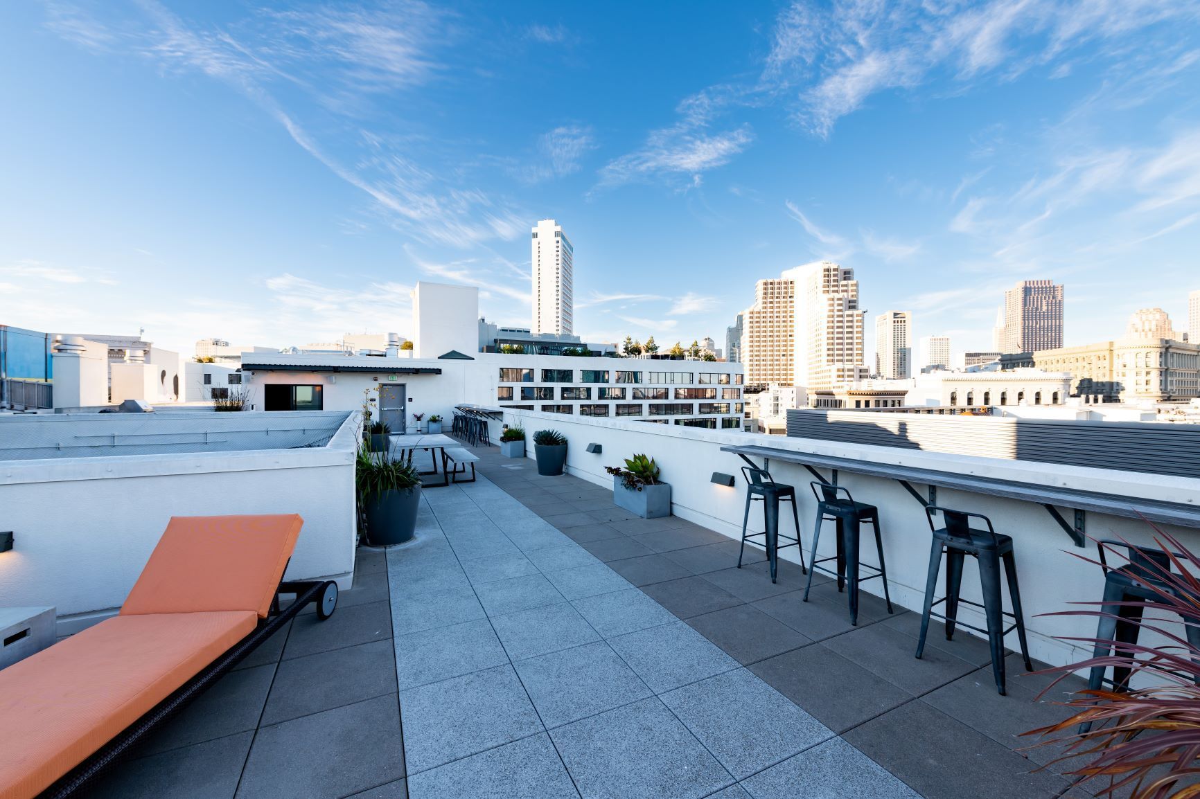 Rooftop patio with orange lounge chairs, bar stools, and city skyline views under a clear sky