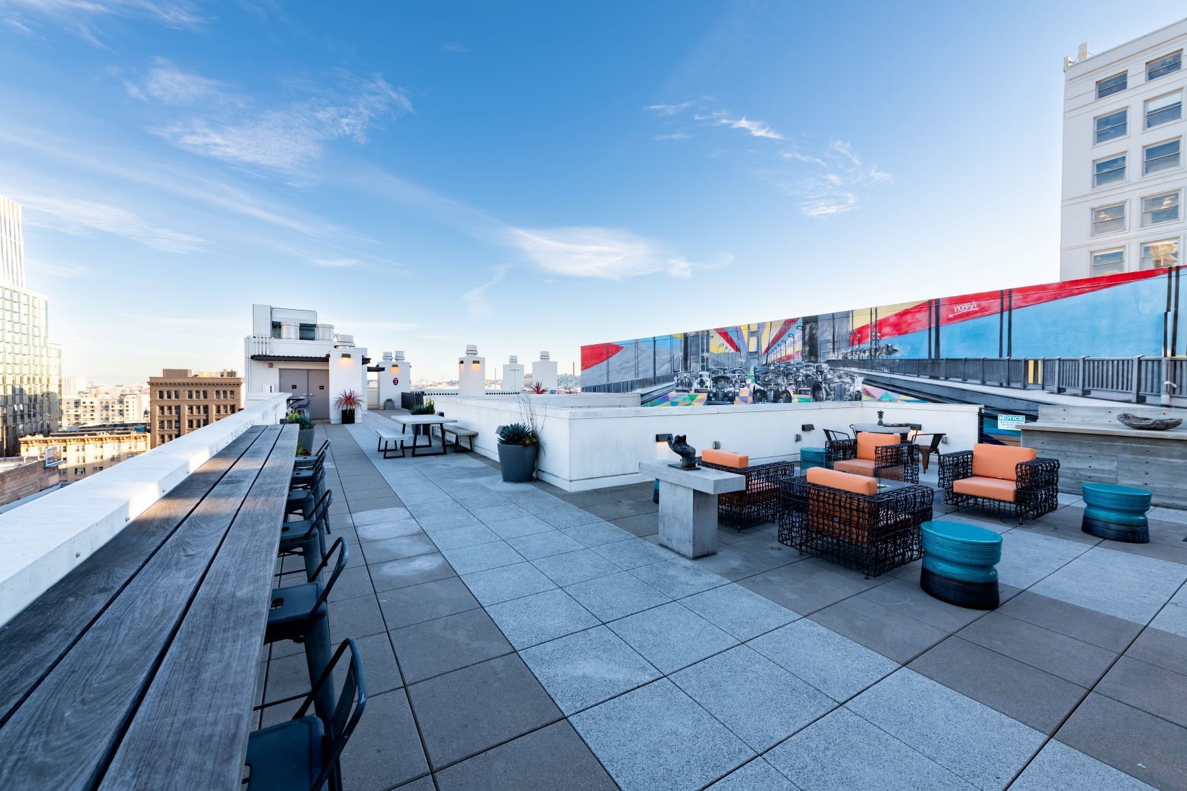 Rooftop terrace with tables, chairs, benches, and city skyline under a blue sky