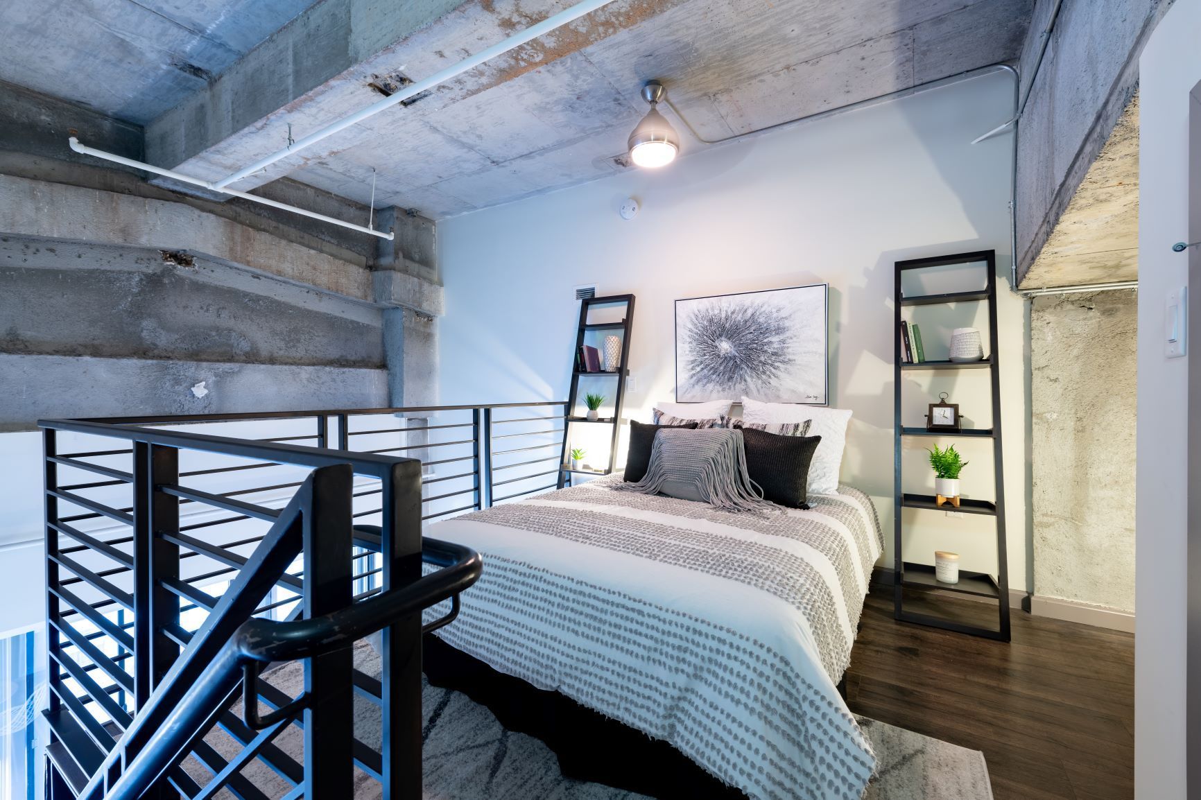 Loft bedroom with a bed, black railings, exposed concrete ceiling, and natural light.