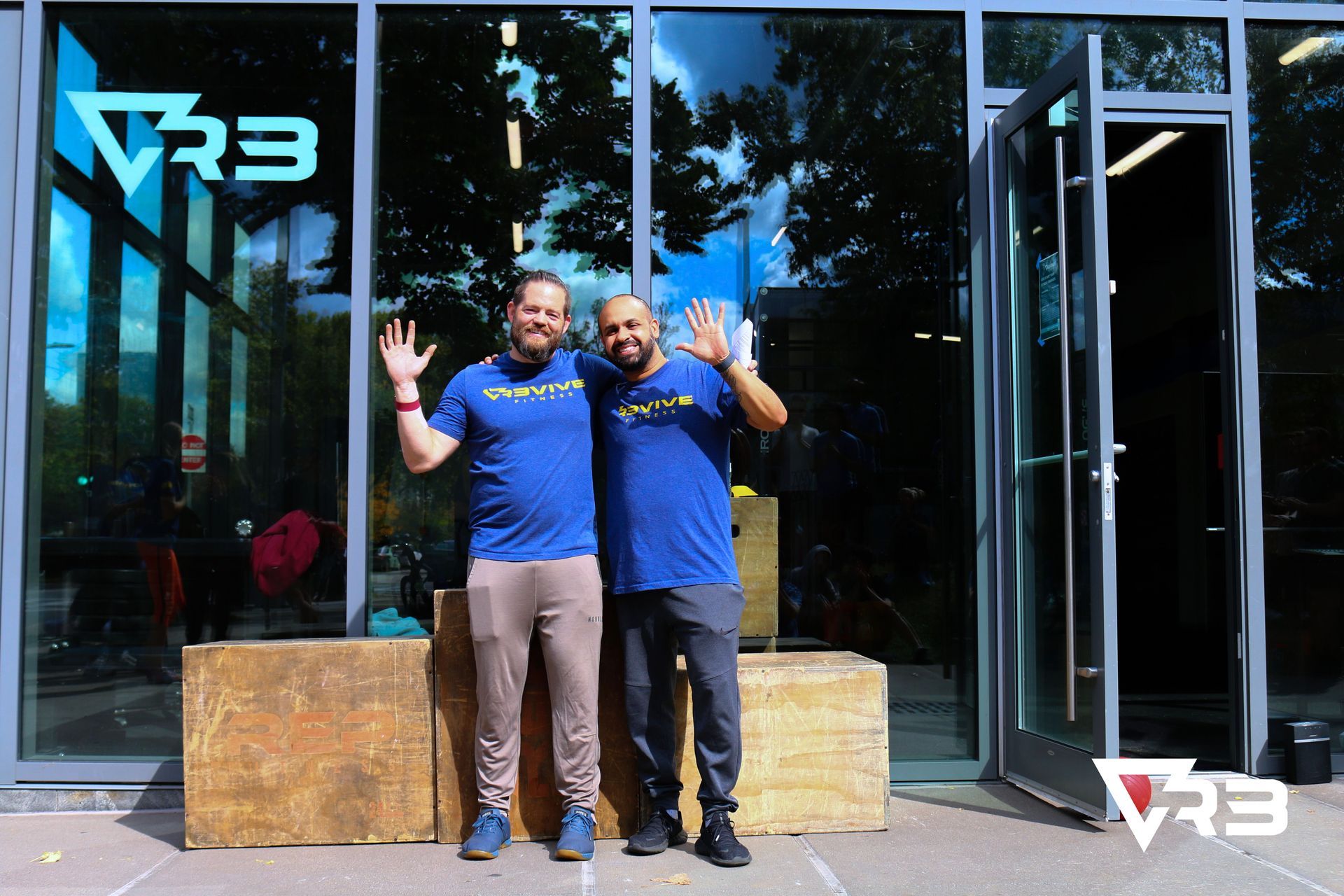 Two men in blue shirts wave in front of a gym with large windows.