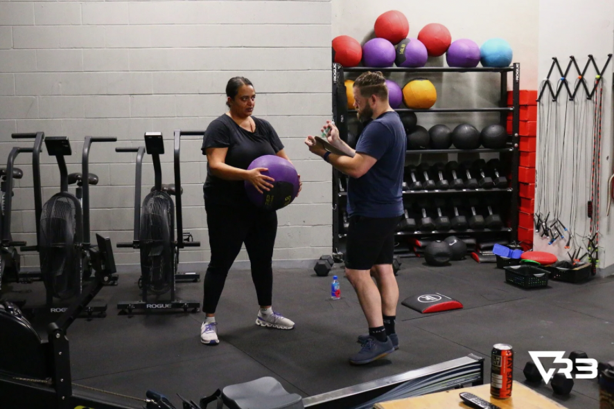 Woman holding medicine ball, trainer instructing in gym.