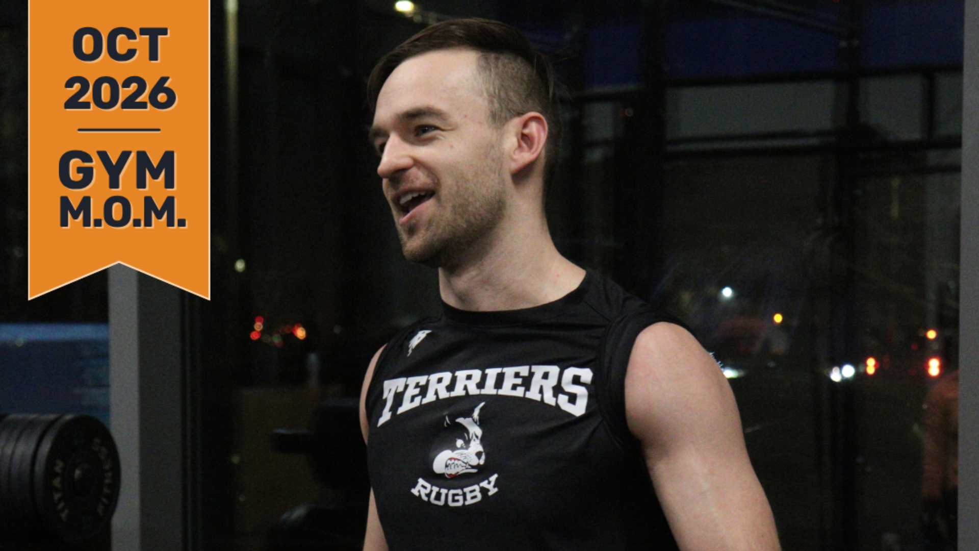 A man smiling inside a gym while holding a weight plate. He is wearing a black sleeveless shirt th