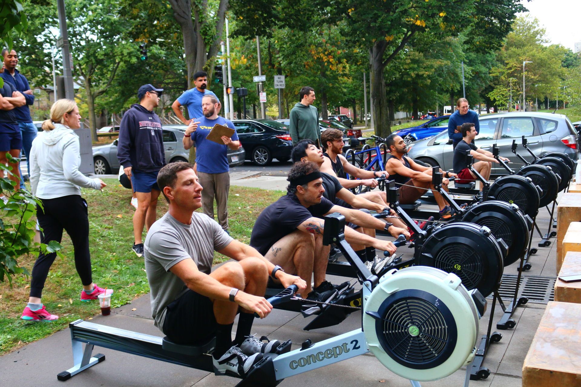 People rowing on machines outdoors, watched by onlookers, in a park setting with cars.
