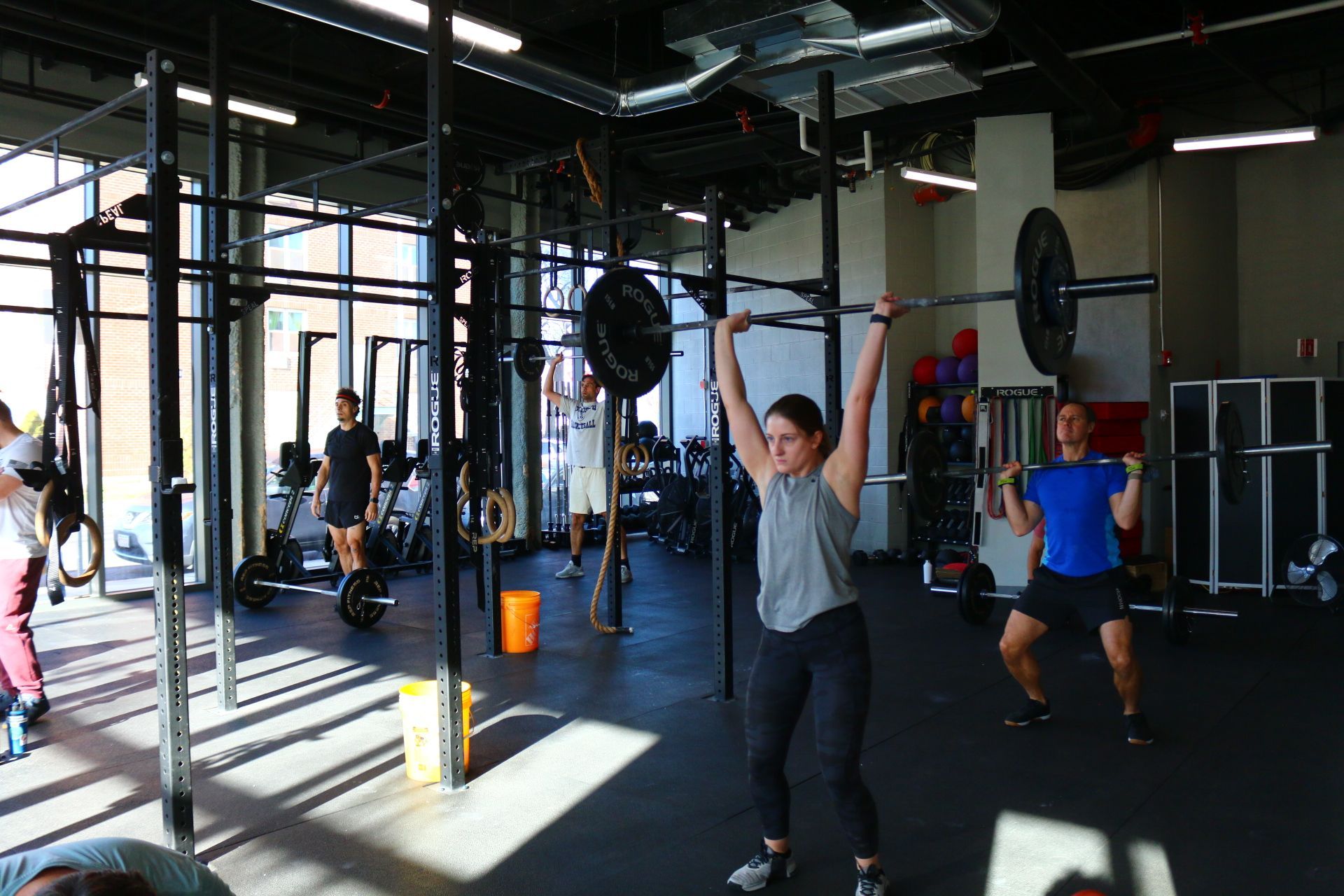 People lifting weights at a gym. Woman overhead presses a barbell, man in blue shirt curls.