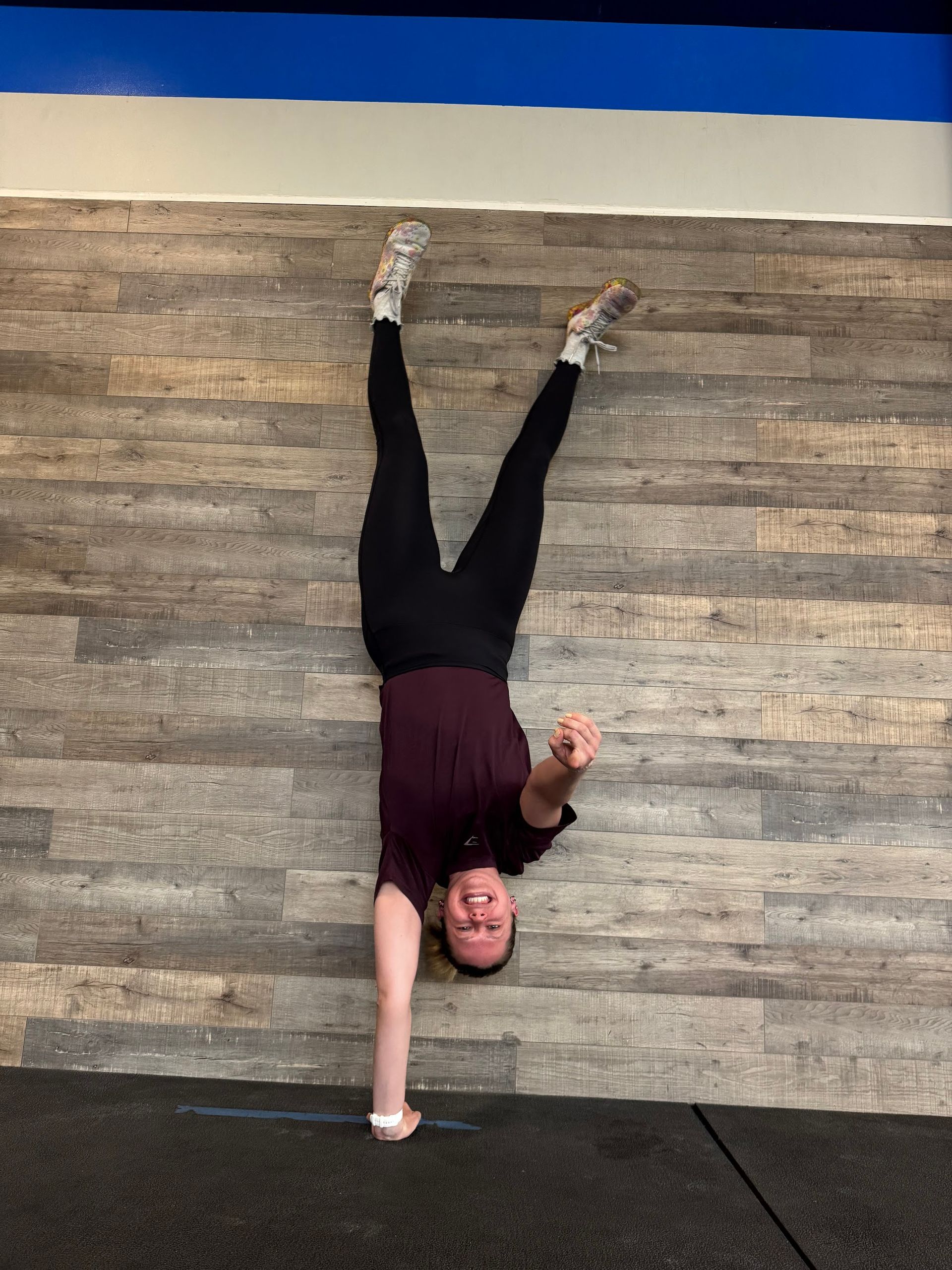Person in a handstand against a wooden wall, smiling. Black pants, maroon shirt.