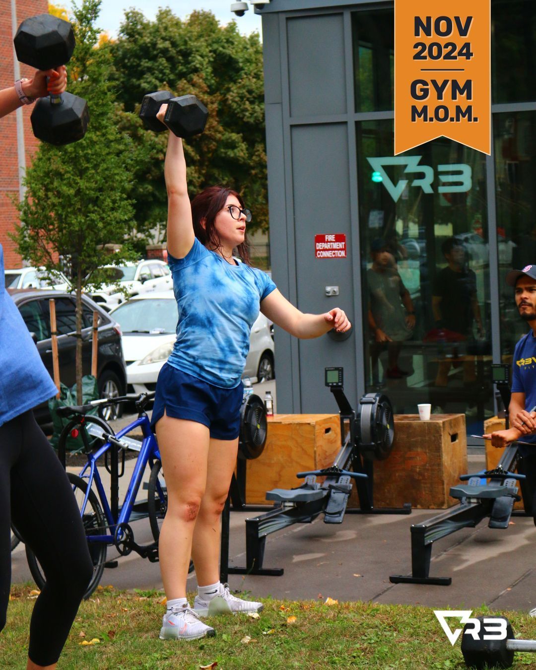 Woman lifting dumbbell overhead at outdoor gym. Blue tie-dye shirt, short shorts, sunny day.