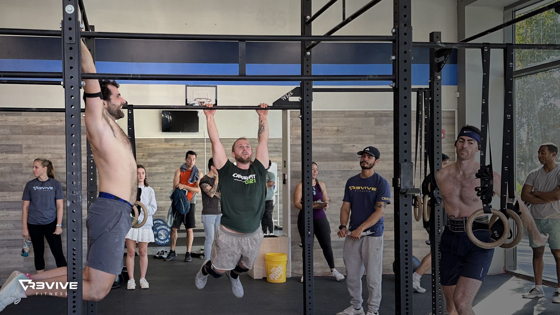 Three men doing pull-ups and ring exercises in a gym. People watching.