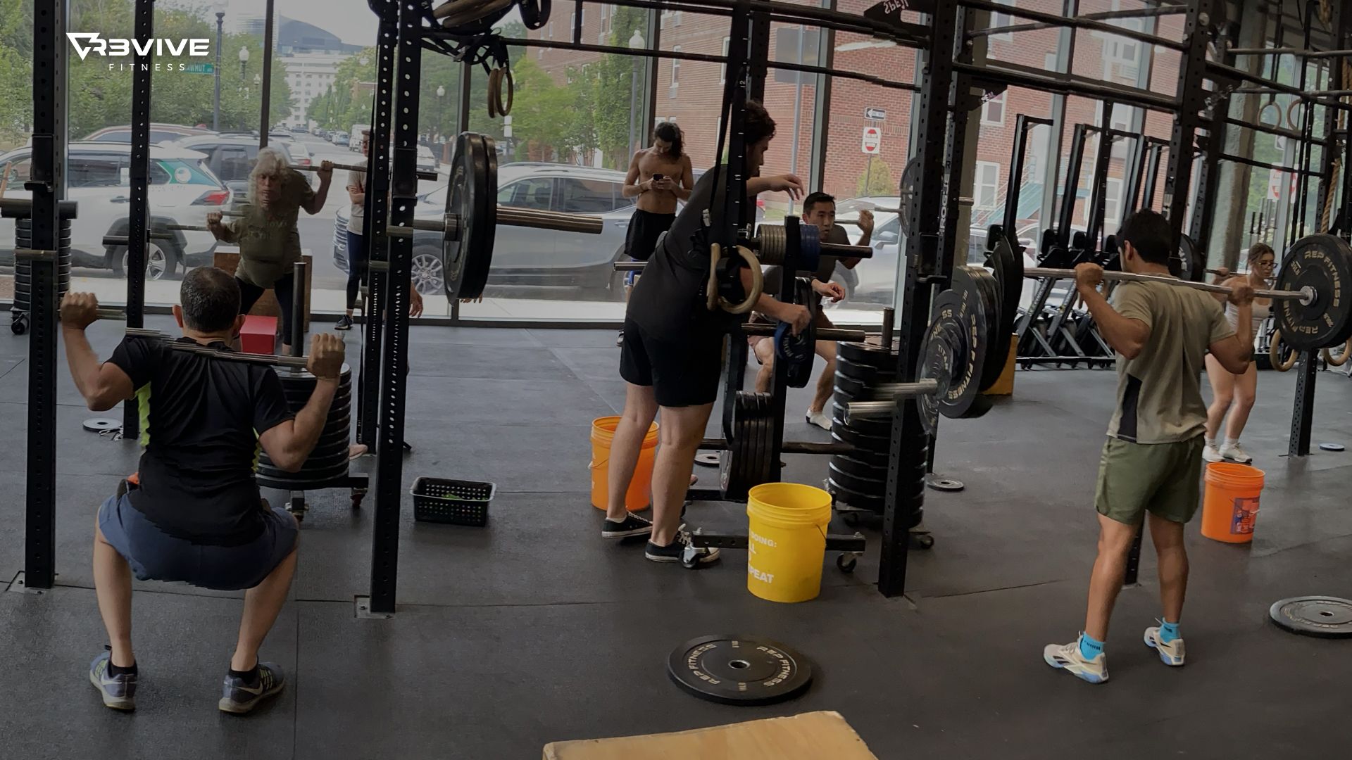 People squatting with weights in a gym. Multiple barbells and weight plates are visible.