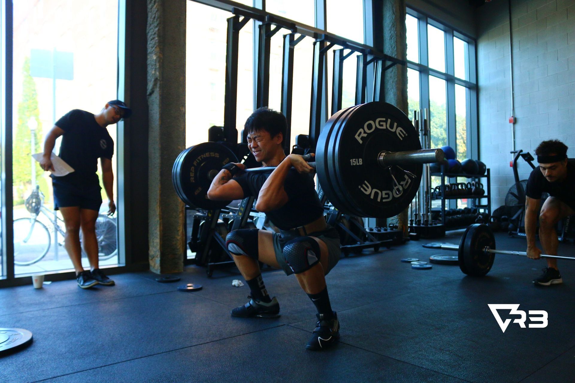 A person squats with a barbell in a gym. Two other people observe.