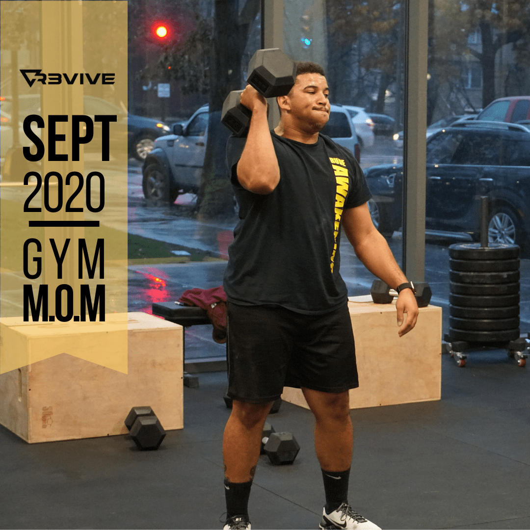 Man lifting a dumbbell in a gym. Black shirt, shorts, and socks. 