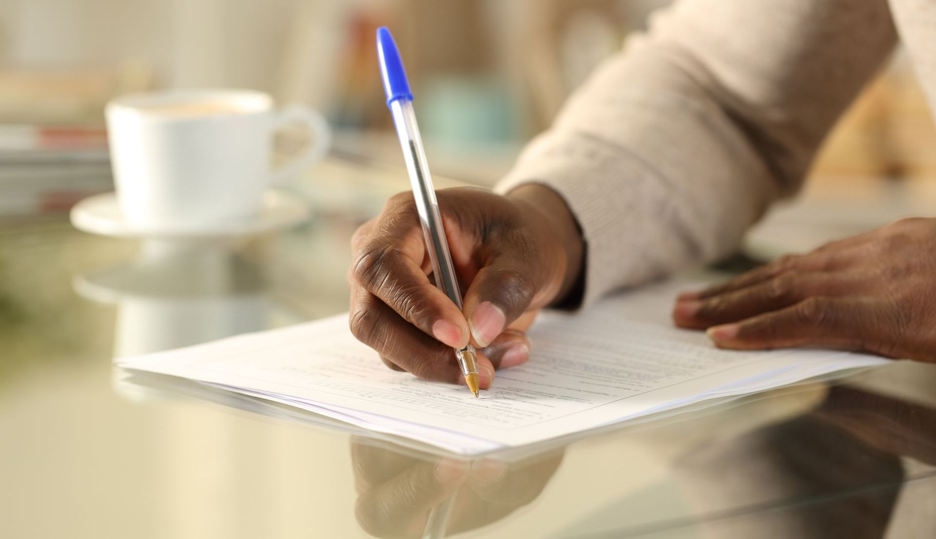 Hand writing on a notebook at a table with a pen, beside a cup of coffee.