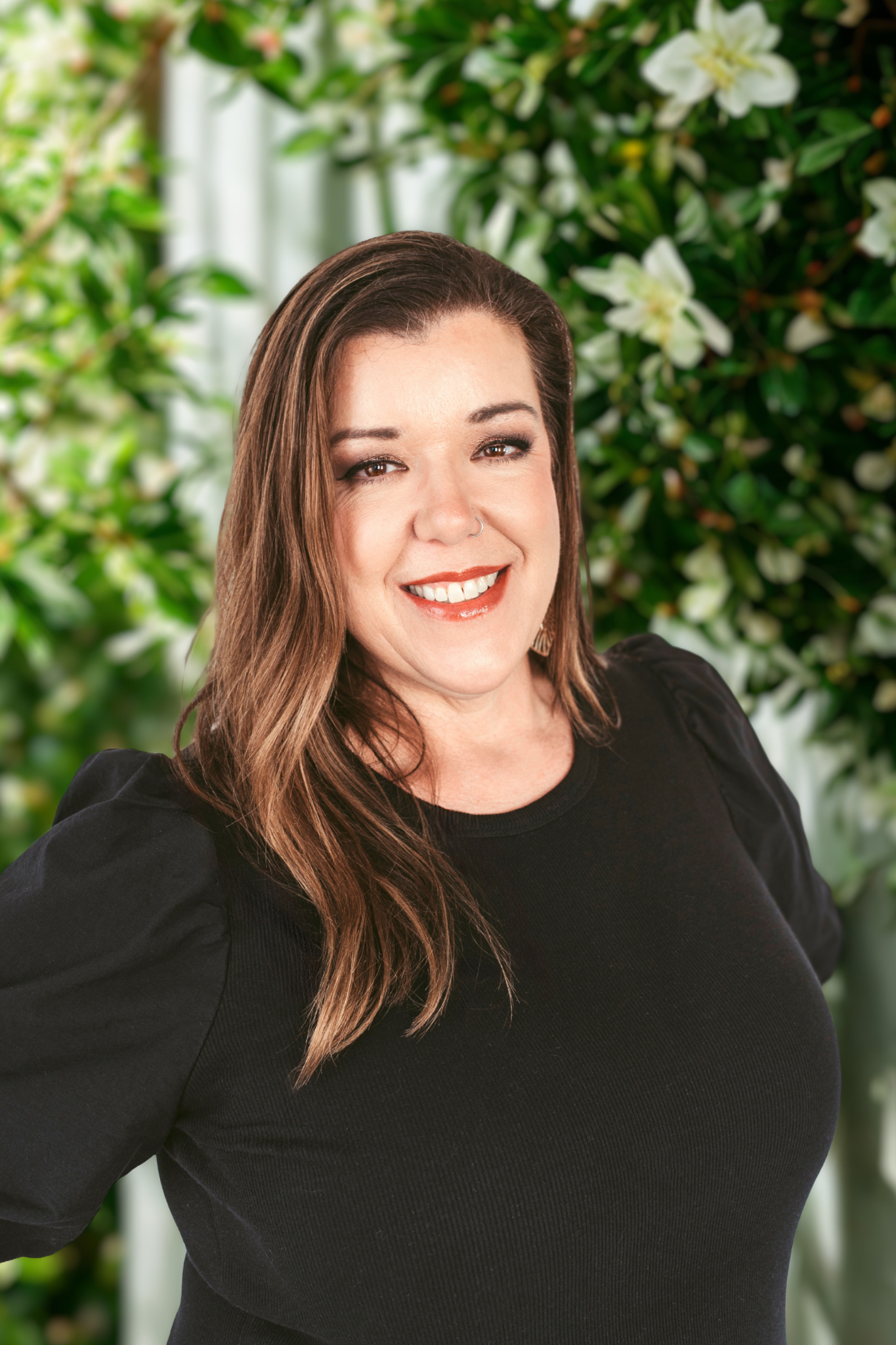 Smiling woman with long hair in a black top, standing before a leafy floral wall.