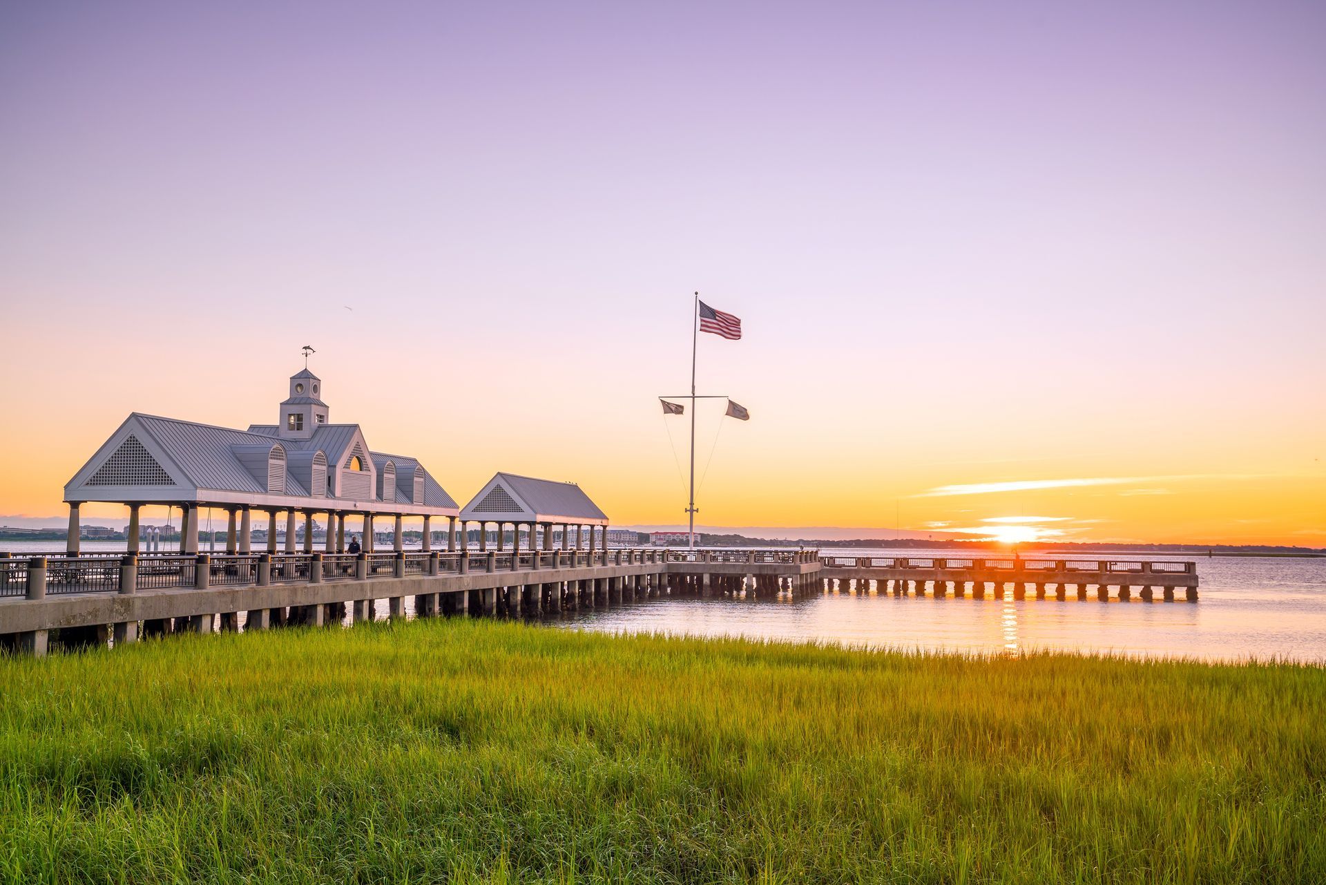 Wooden pier with covered sections, American flag, grass in foreground, and sunset.