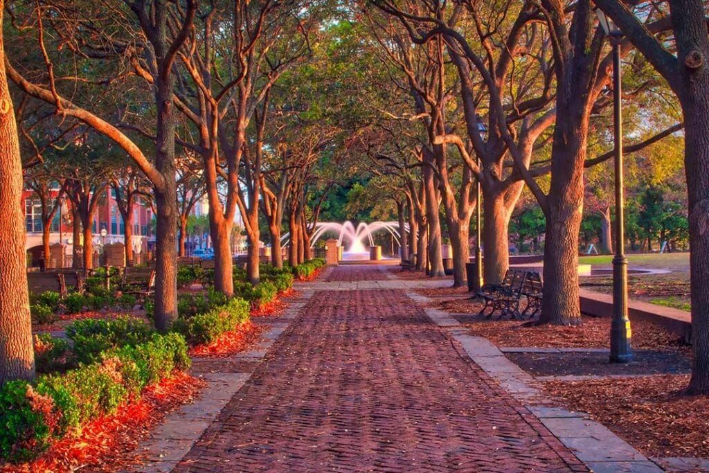 Brick path lined with trees and manicured bushes, leading to a fountain under an archway.