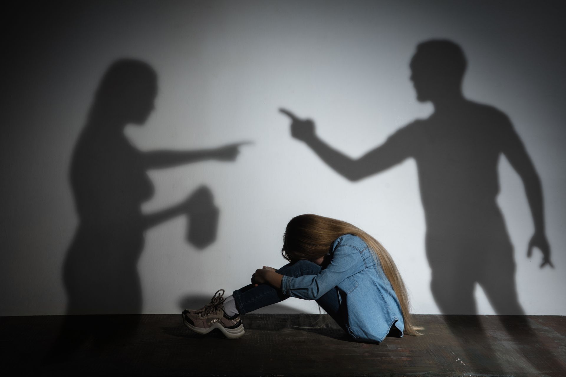 A person sits on the floor, head in knees, shadows of two people pointing at them representing dv